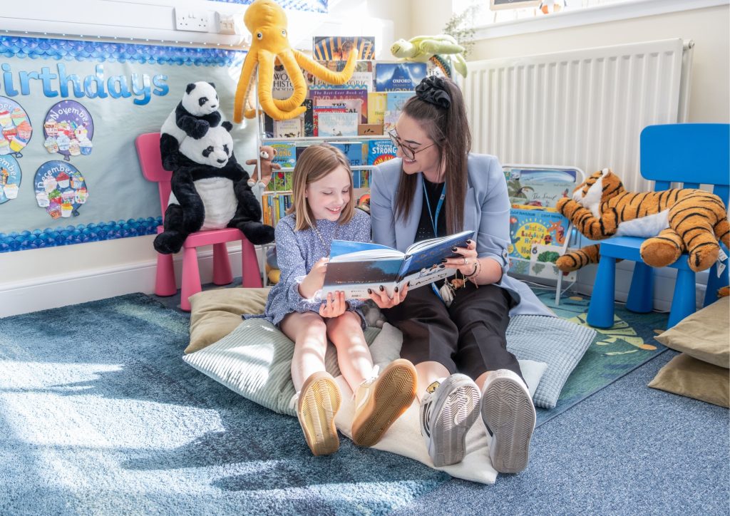 Lydia and a student smile, while reading a book in the library corner of the learning spacec